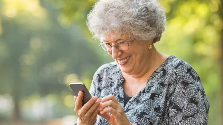 Mulher idosa sorrindo ao olhar o celular em um quintal ensolarado, feliz com os resultados de ganhar dinheiro online de forma simples e segura.