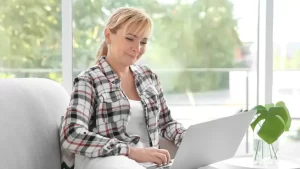 Mulher de 50+ sentada com o laptop no colo em ambiente tranquilo, representando leveza e acolhimento para quem tem dúvidas sobre começar no digital.